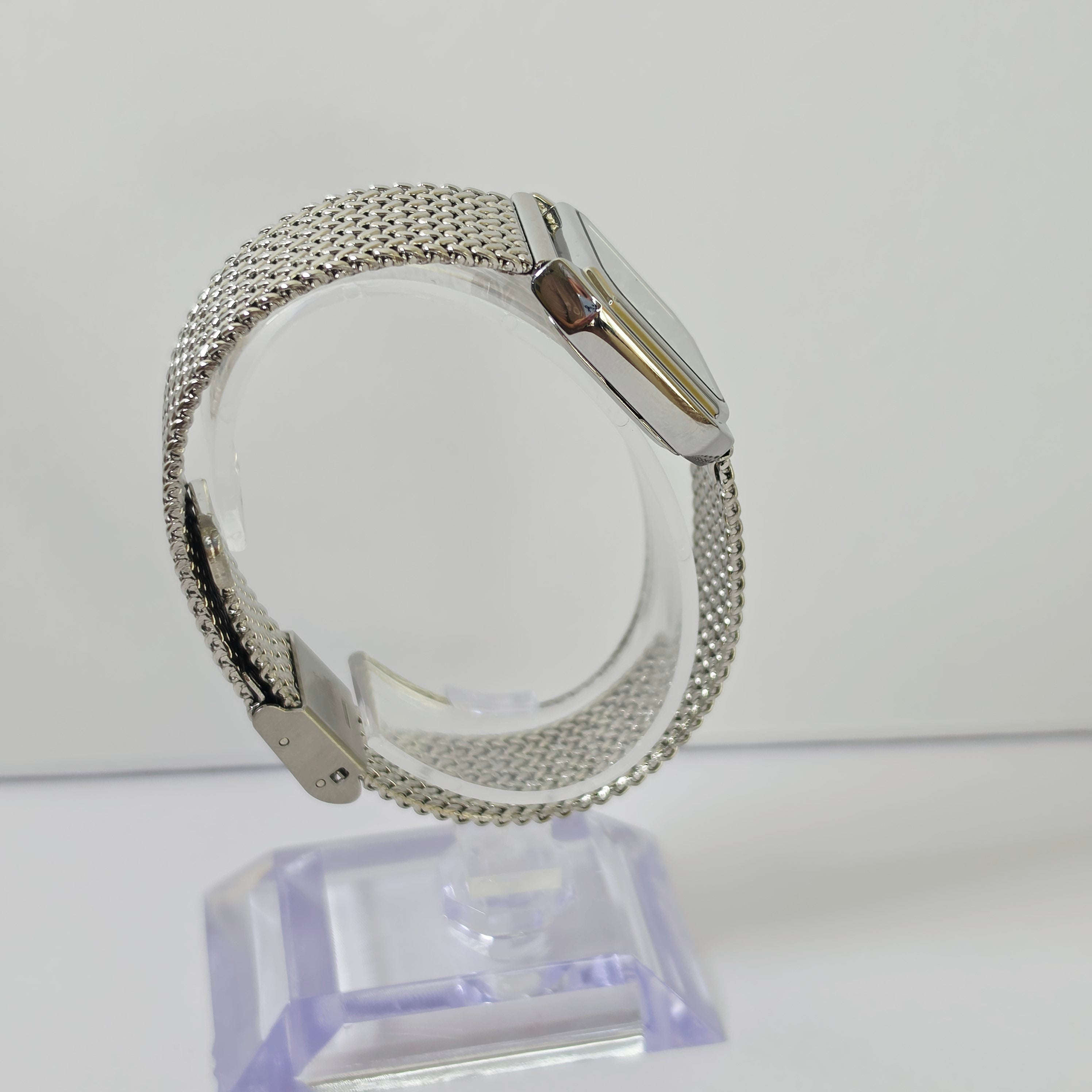 A silver-tone mesh watch band with a matching silver watch case, displayed on a clear acrylic stand against a plain white background.