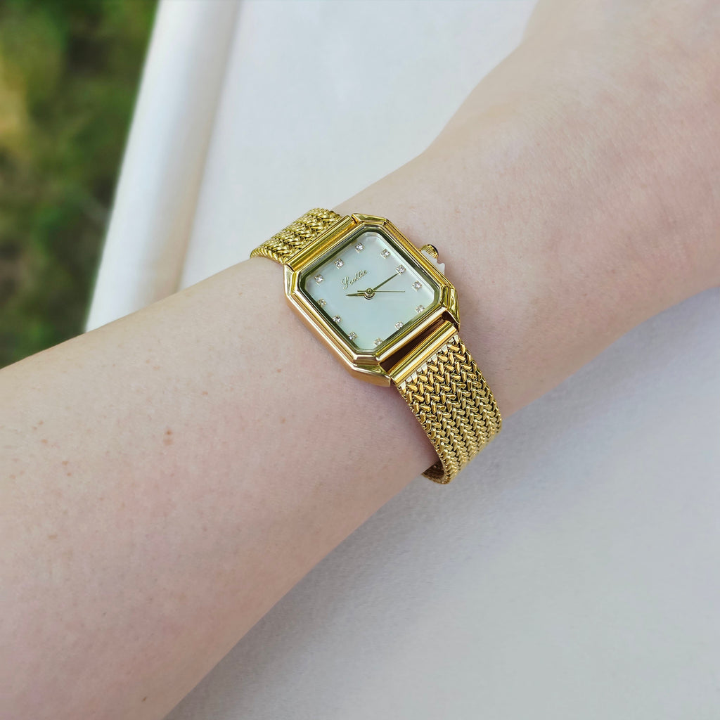 A gold-tone rectangular mesh watch with a white dial, worn on a person's wrist against a light neutral background.