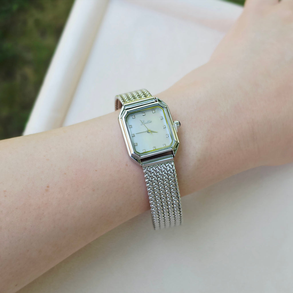 A silver-toned women's watch with a rectangular bezel and mesh strap, worn on a wrist. The watch has a white dial with gold-tone hands and crystal rhinestone hour markers.