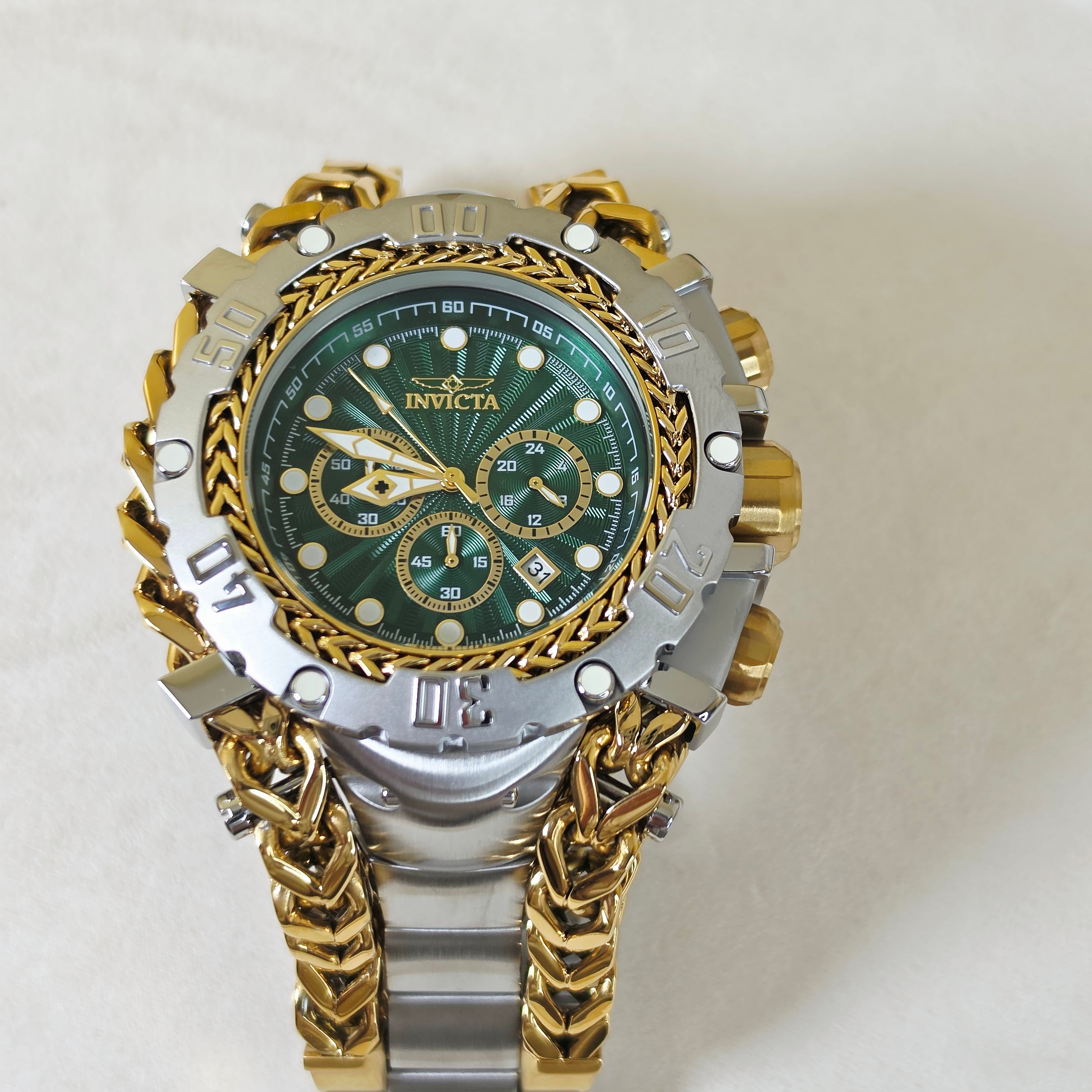 An Invicta wristwatch with a green dial, a two-tone silver and gold-tone case, and a chunky gold-tone chain link bracelet, photographed against a light beige background.