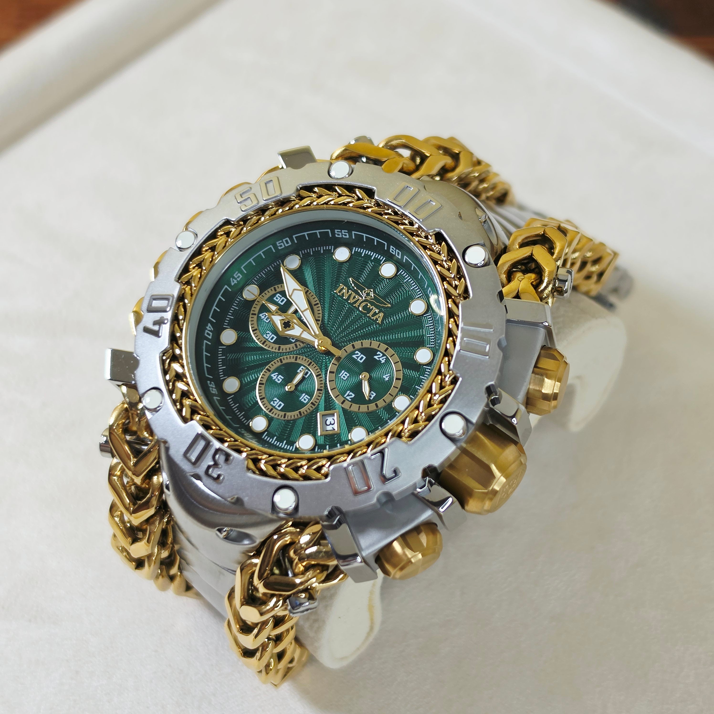 An Invicta wristwatch with a green sunburst dial, silver and gold-tone cases, and a gold-tone chain bracelet, displayed on a white surface.