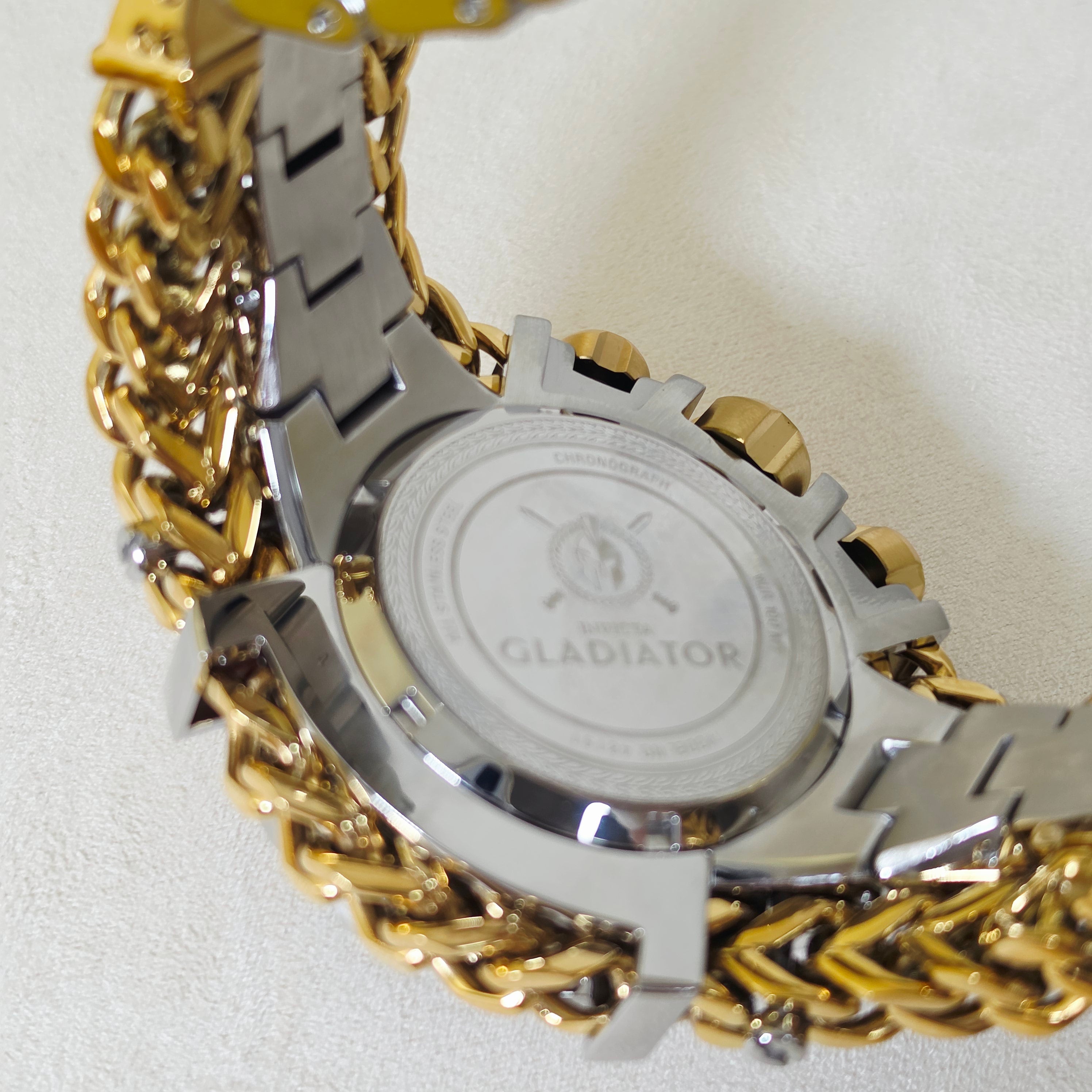 Close-up of the back of a Gladiator watch with a two-tone silver and gold-tone chain link bracelet on a light textured background.