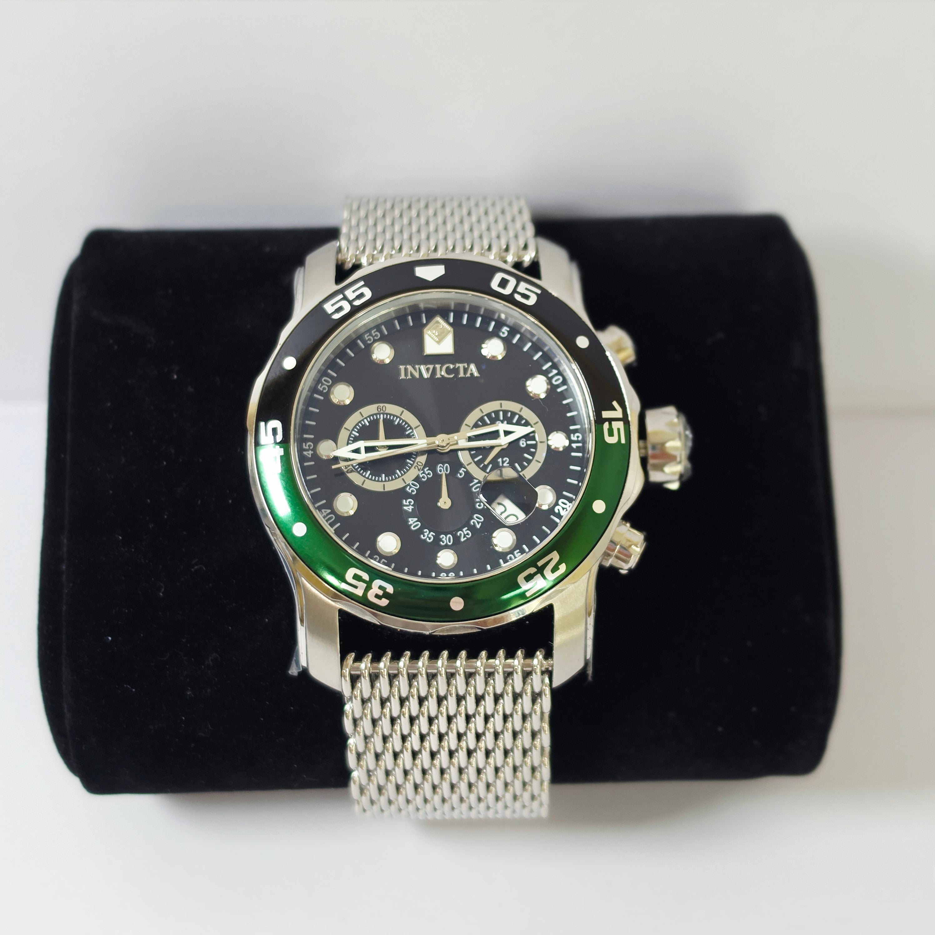 An Invicta wristwatch with a silver mesh strap, black dial, and green bezel, displayed on a black velvet cushion against a white background.
