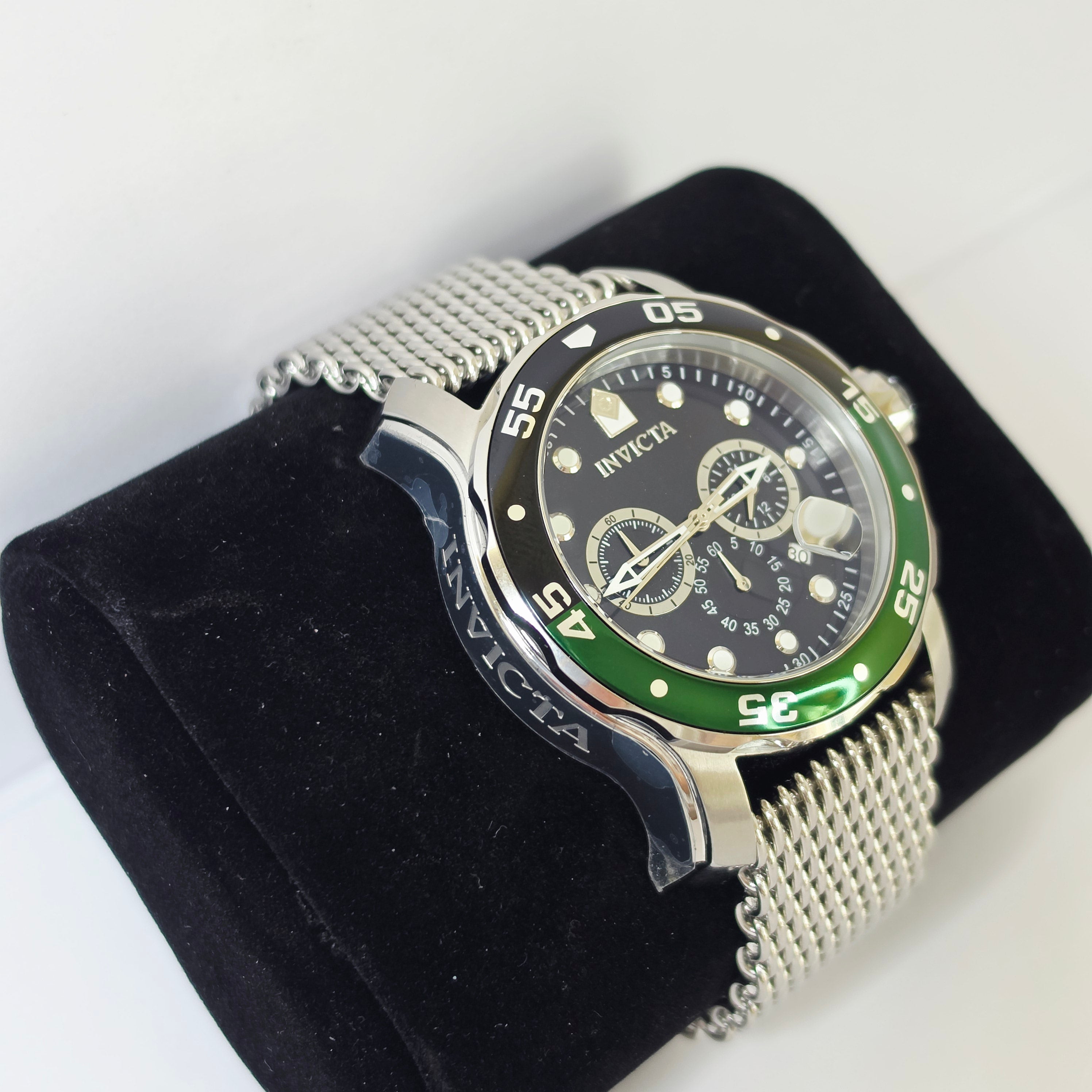 An Invicta wristwatch with a silver mesh bracelet, black dial, and a green and black bezel, displayed on a black velvet watch cushion against a white background.
