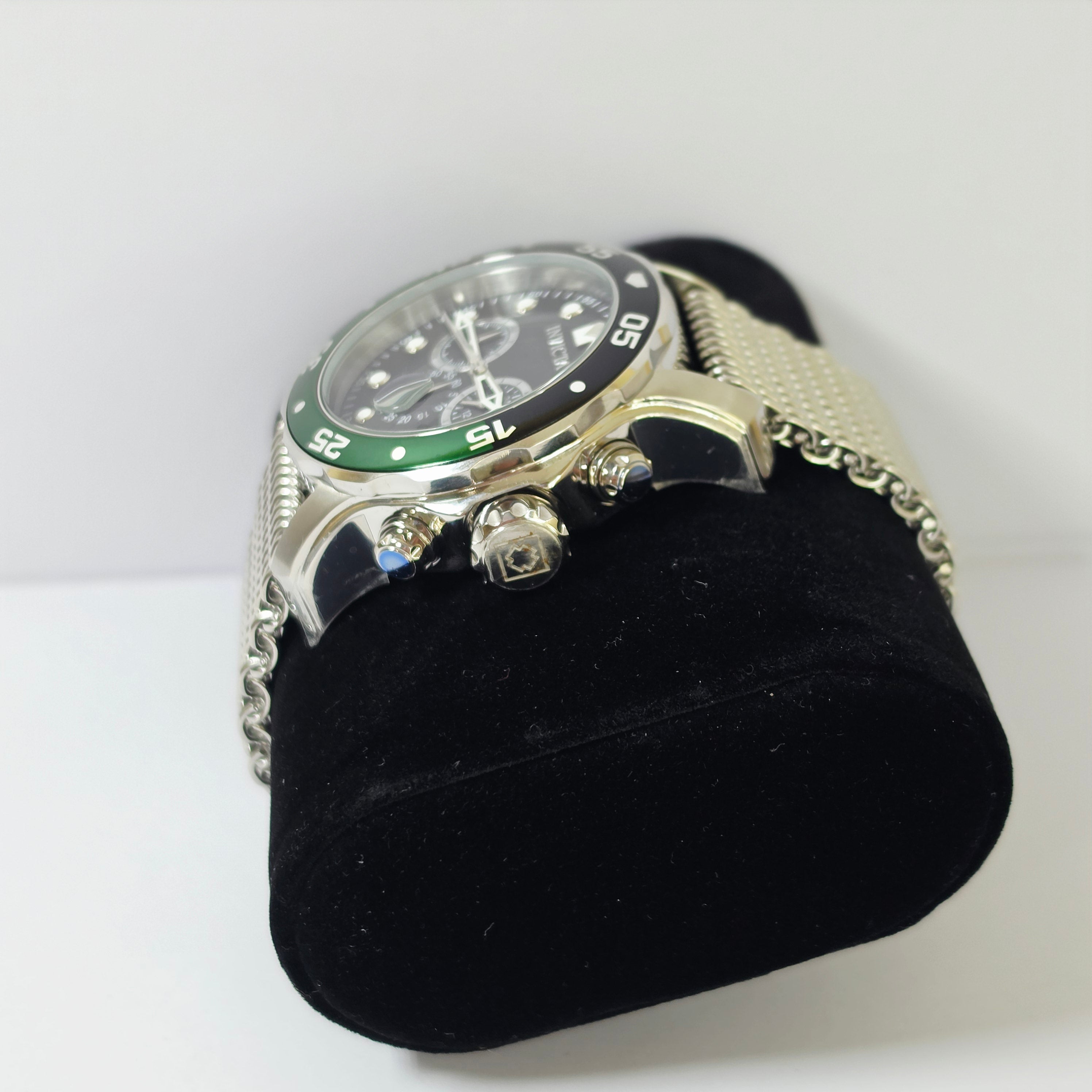 A silver-toned stainless steel watch with a black dial, green bezel insert, and mesh metal bracelet, displayed on a black velvet wristlet stand against a white background.