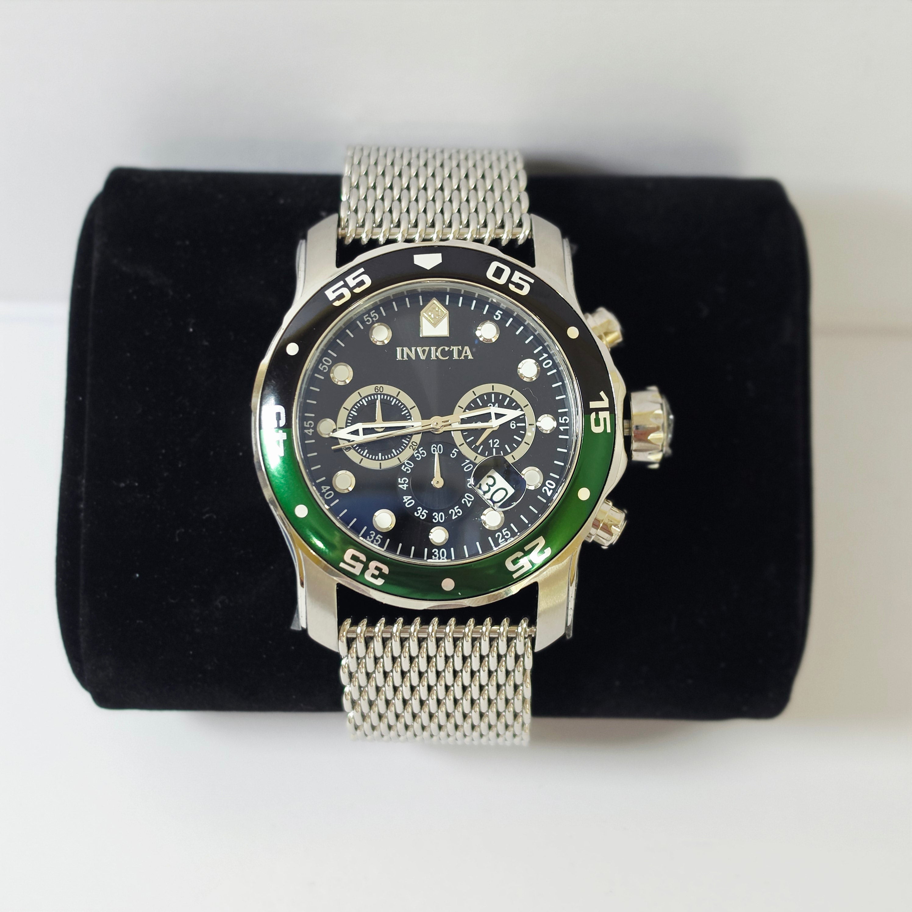 An Invicta stainless steel wristwatch with a black dial, green bezel, and silver mesh strap, displayed on a black velvet watch cushion against a white background.