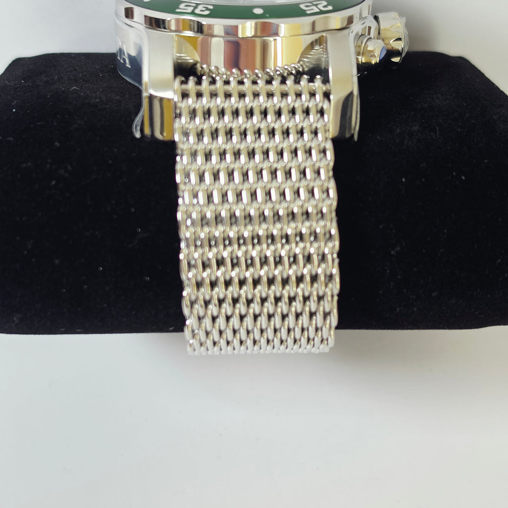 A close-up of a silver-toned metal mesh watch band attached to a watch case with a green dial, displayed on a black velvet stand against a white background.
