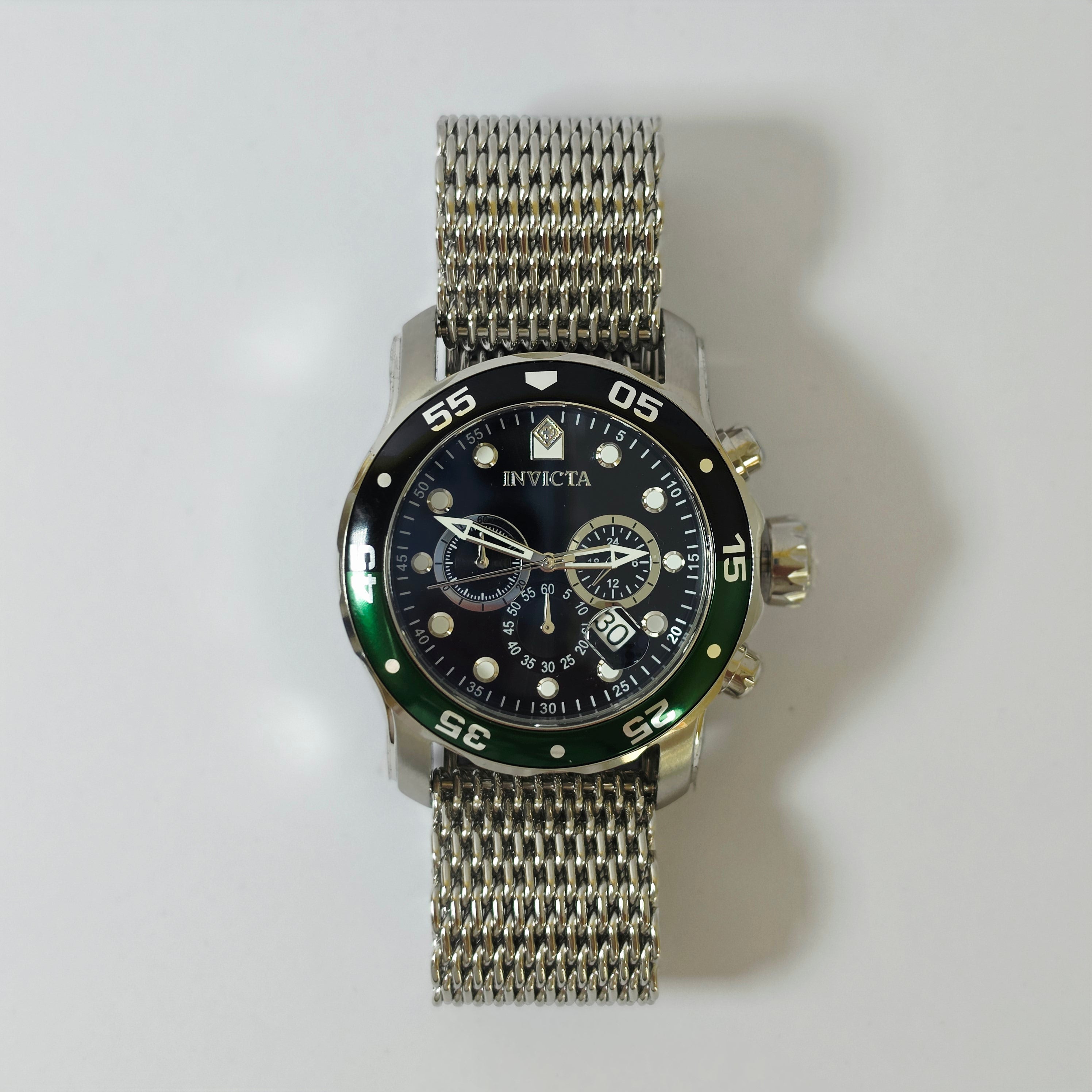 An Invicta wristwatch with a silver mesh metal bracelet, a black dial, and a black bezel with a green accent, photographed against a light gray background.