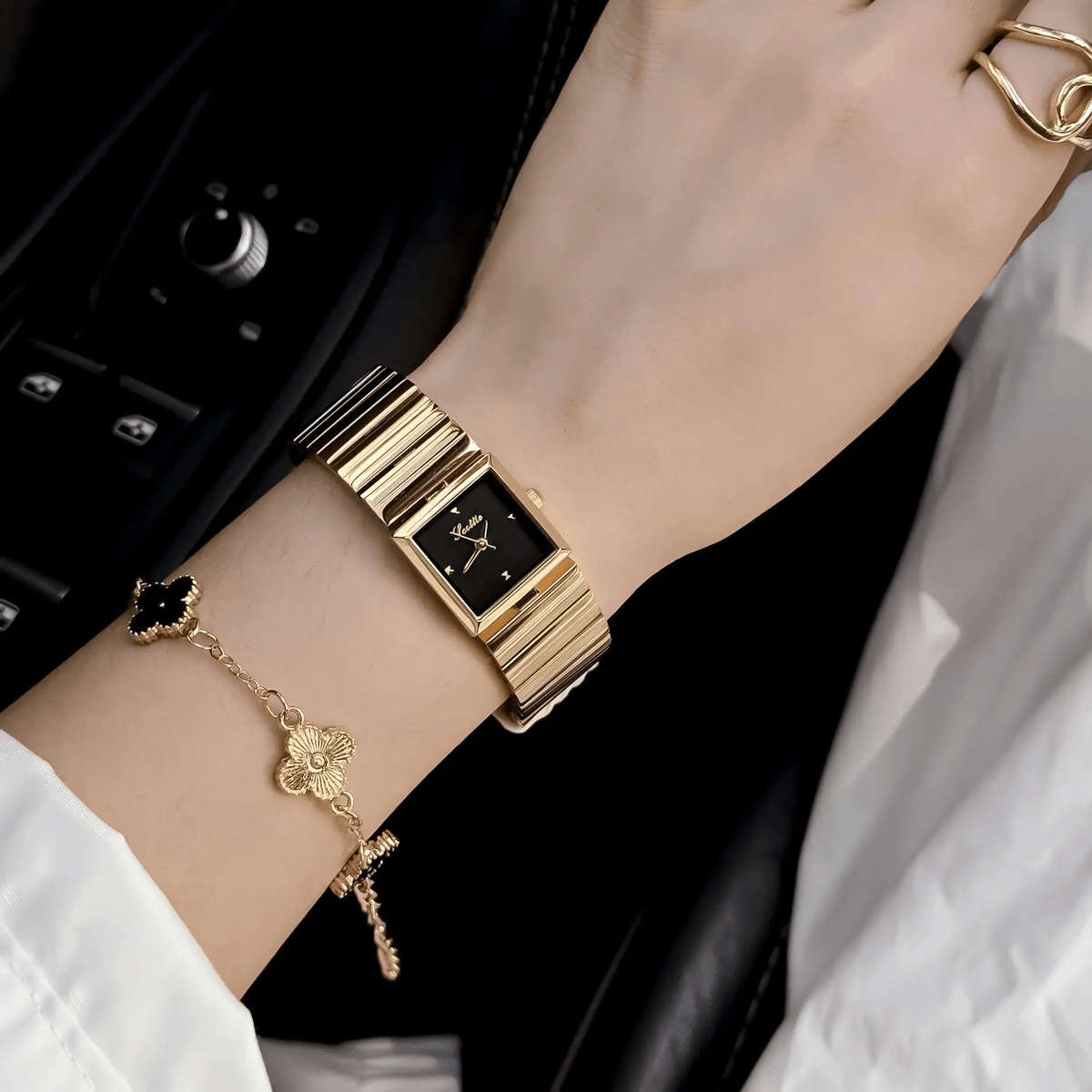 Black dial gold square bracelet watch worn on wrist with layered bracelets
